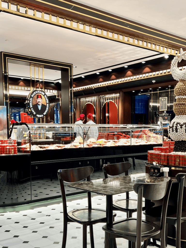Interior of the Hafız Mustafa dessert shop at Galataport Istanbul, with elegant black and gold decor, rows of sweets behind a glass counter, and staff in red hats (called fez) preparing orders.