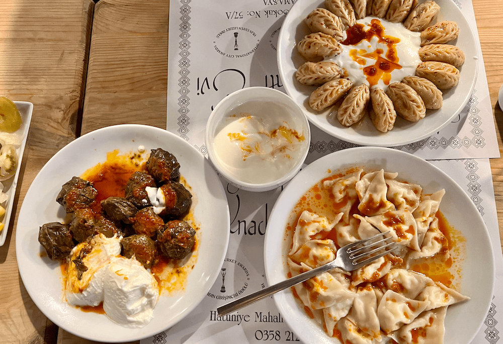 A variety of Amasya specialties including stuffed grape leaves with yogurt, two types of mantı (Turkish dumplings) with yogurt and butter sauce, served on a wooden table.