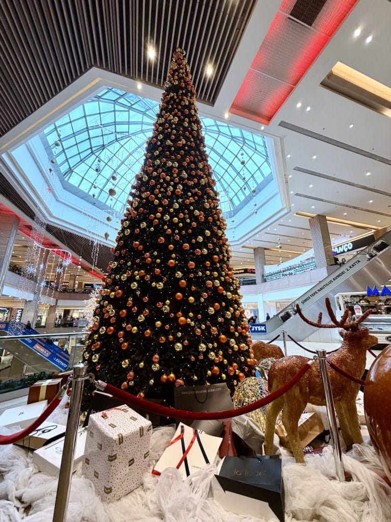 Giant Christmas tree decorated with red and gold ornaments inside a modern Istanbul shopping mall, surrounded by wrapped presents and reindeer figures.