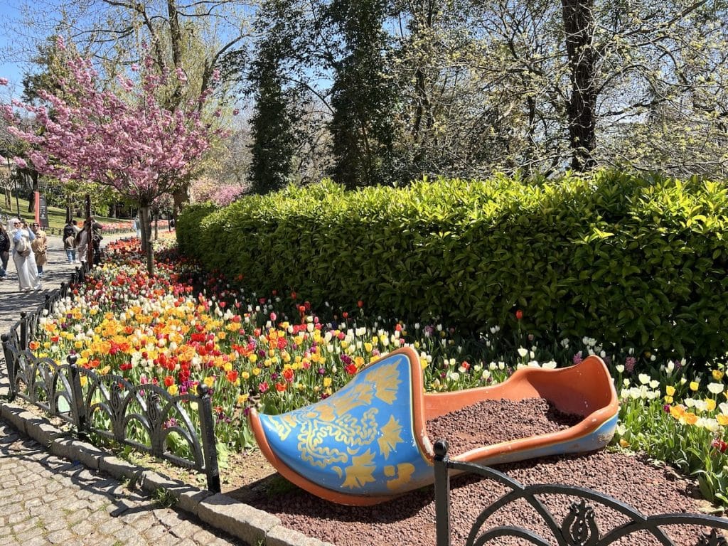 A giant ceramic tulip shoe sculpture in Emirgan Park displayed beside a walkway lined with multi-colored tulips and flowering trees.