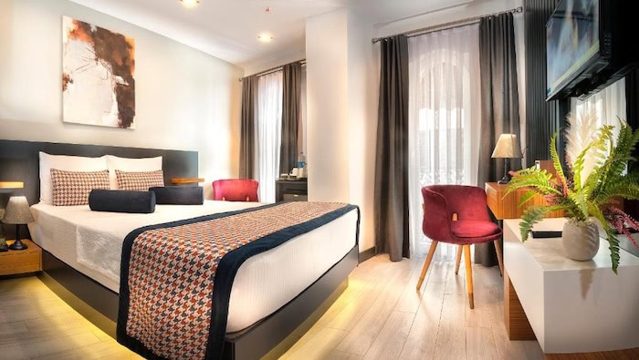 Cozy, modern hotel room in Istanbul with a neatly made bed, desk, maroon accent chair, and large windows with flowing curtains.