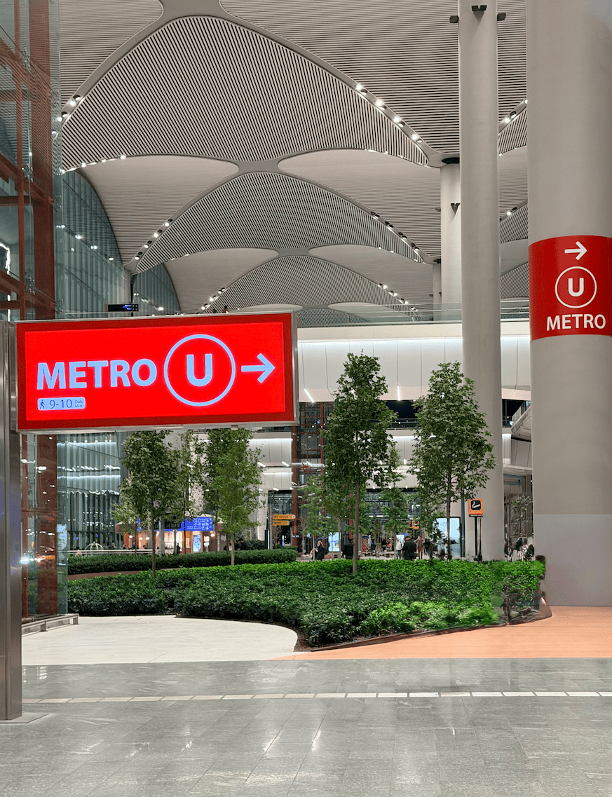 Red airport sign reading "METRO U" with an arrow pointing toward the metro station inside Istanbul Airport showing a public transport option 